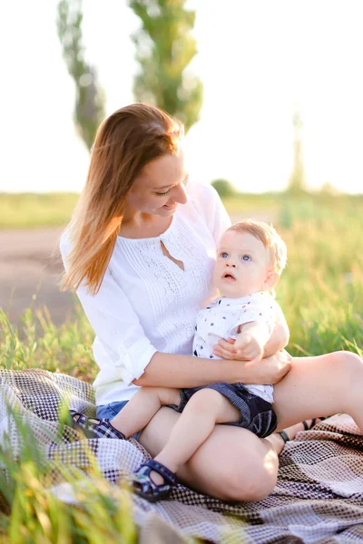 Young caucasian mother sitting with little child on plaid, grass on background. — Stock Photo, Image