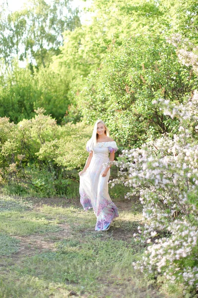 Jonge vrouw dragen witte jurk staande in de buurt van witte lila bloemen. — Stockfoto