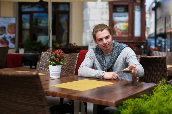 Hombre joven sentado en la cafetería de la calle y usando suéter gris . — Foto de Stock