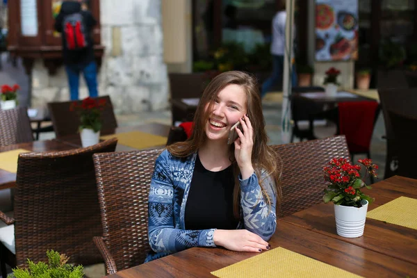Chica joven hablando por teléfono inteligente en la cafetería cerca de flores rojas . — Foto de Stock