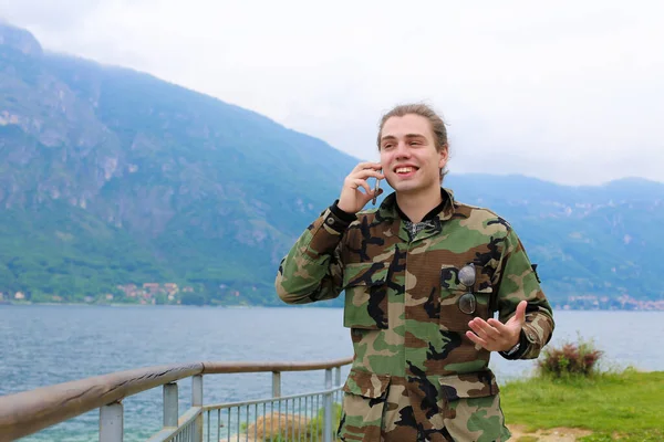 Młody człowiek uśmiechający się rozmawia przez smartfon w pobliżu poręczy, Jezioro Como i Alpy góry w tle. — Zdjęcie stockowe