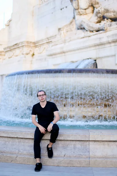 Joven hombre de negocios sentado cerca de la fuente de Trevi, con gafas y ropa negra . — Foto de Stock