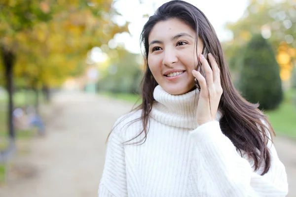 Asiática bonita chica vistiendo suéter blanco hablando por teléfono inteligente y caminando en parque . — Foto de Stock