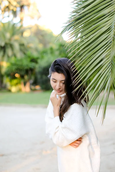 Korean dreaming girl in white sweater standing near palm leaf. — Stock Photo, Image