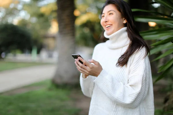 Enfoque en el teléfono inteligente en manos de mujer china, chica de pie en el parque tropical . — Foto de Stock