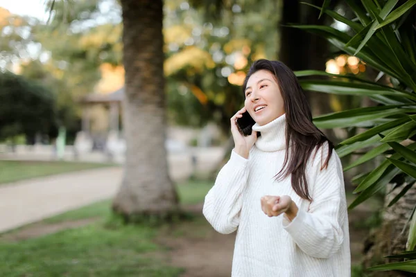 Chica asiática en parque tropical hablando por teléfono inteligente cerca de la palma . — Foto de Stock