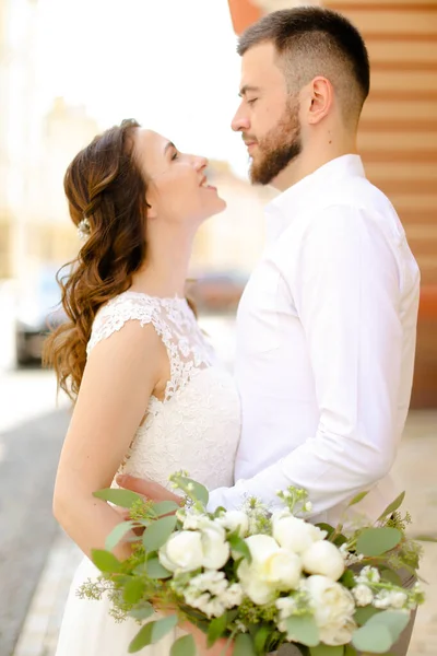 Feliz noivo abraçando noiva mantendo flores e vestindo vestido branco . — Fotografia de Stock