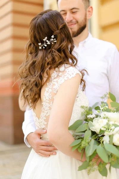 Feliz noivo sorridente abraçando noiva mantendo flores e vestindo vestido branco . — Fotografia de Stock