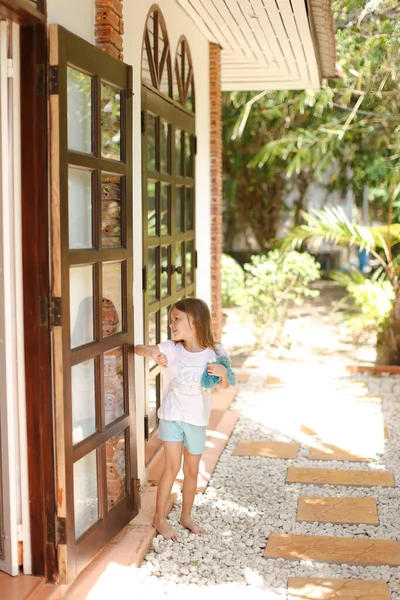 Pequeña chica descalza parada cerca de la puerta con juguete afuera, palmas en el fondo . — Foto de Stock