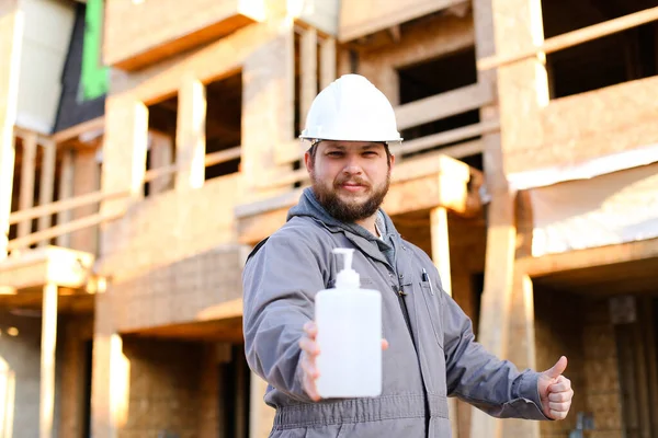 Bauarbeiter in Harthut zeigt Desinfektionsmittel und Daumen hoch auf Baustelle. — Stockfoto