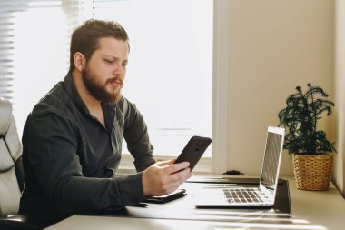 Ofiste çalışan erkek yönetici smatrphone ve laptop kullanıyor.