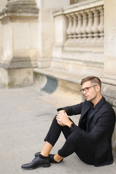 Young stylish man sitting on sidewalk ground and wearing black suit.