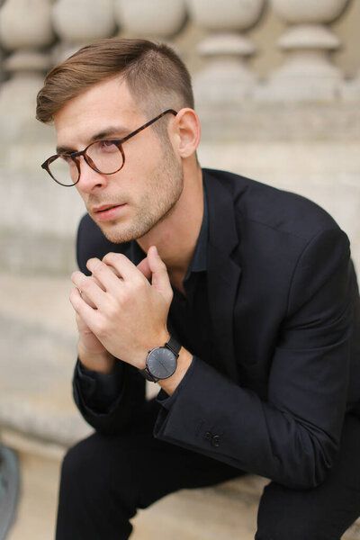 Youn handsome male person sitting on sidewalk leaning on concrete banister.