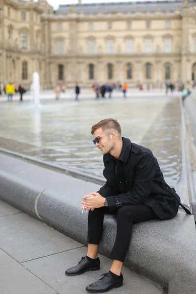 Young man wearing black suit sitting near fountain and smoking in Paris, France. — Stock Photo, Image