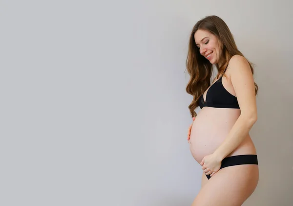 Siyah iç çamaşırlı hamile bir kadın. Gri fotokopi arkaplanda karnını tutuyor.. — Stok fotoğraf