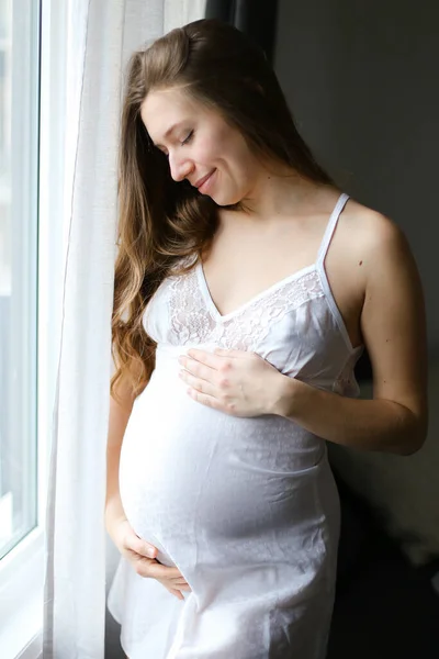 Genç, güzel, hamile bir kadın beyaz elbiseli, pencerede duruyor.. — Stok fotoğraf