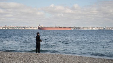 Çakıl taşlı sahilde bir balıkçıyla boğaz kenarındaki Elsinore, Danimarka 'da bir kargo gemisini oveloklayan ses, Ağustos 2025