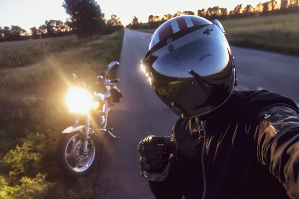 一个男人在头盔上对着摩托车在路上的傍晚 — 图库照片