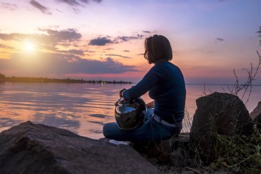 Bir motosiklet kask bir kızla günbatımı çerçevede bir nehrin kenarında oturur