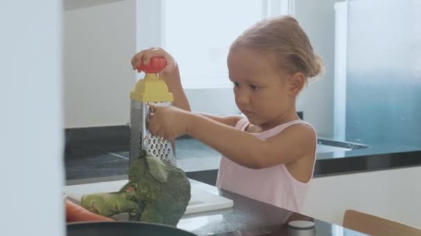 Mignon petit enfant fille frotter la carotte sur une râpe à la cuisine domestique . — Video