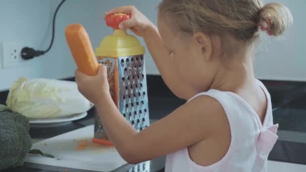 Mignon petit enfant fille frotter la carotte sur une râpe à la cuisine domestique . — Video