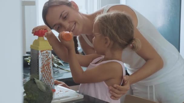 Little girl rubbing carrot on a grater, bits and eats it at domestic kitchen. — Stock Video