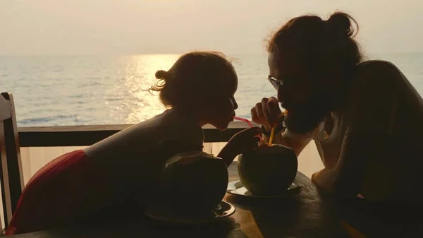 Отец и дочь сидят в кафе с видом на море и пьют кокосы вместе — стоковое фото