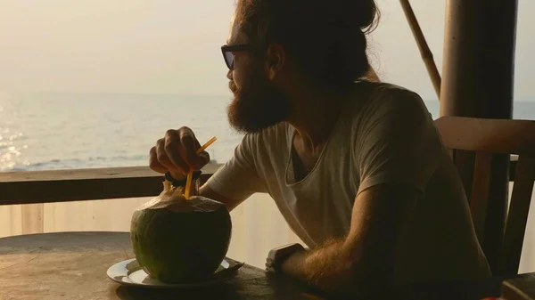 El hombre barbudo se sienta en la cafetería con vistas al mar y bebe agua de coco — Foto de Stock