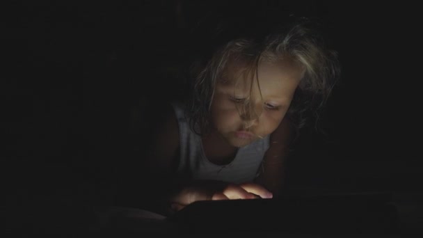 Malá roztomilá holka ležící na posteli a pomocí digitálního tablet zařízení před spaním. — Stock video