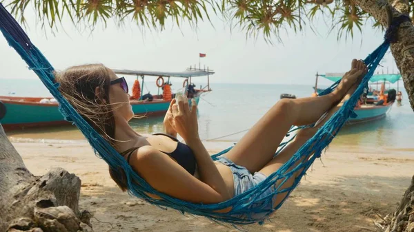 Mujer joven acostada en una hamaca con teléfono inteligente en la playa de arena — Foto de Stock