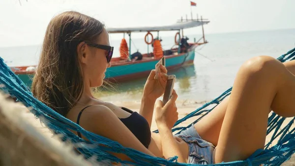 Mujer joven acostada en una hamaca con teléfono inteligente y tarjeta bancaria en la playa de arena — Foto de Stock