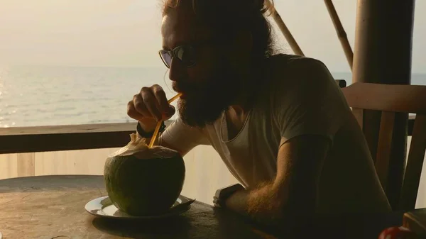 Homem barbudo senta-se no café seaview e bebe água de coco — Fotografia de Stock