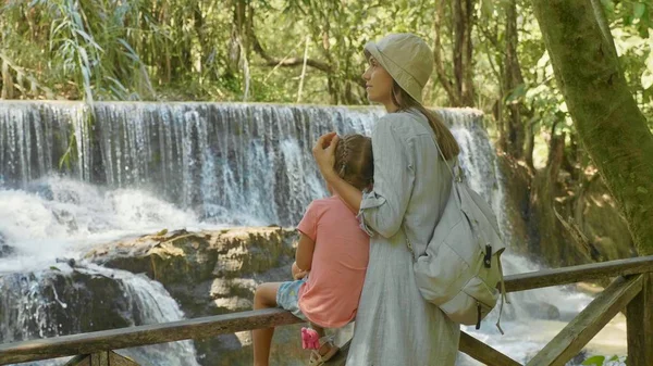 Uma jovem mãe e sua linda filha descansando contra a cascata de uma cachoeira — Fotografia de Stock