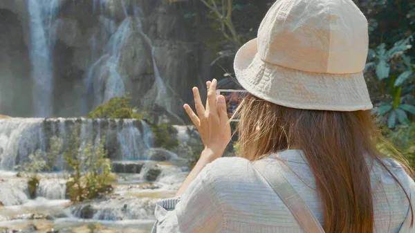 Uma jovem mulher de pé contra a cachoeira e tirar fotos no telefone inteligente — Fotografia de Stock