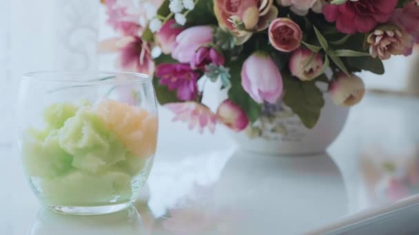 Cuenco de vidrio con exfoliante colorido en el interior y flores en el fondo en el salón de spa — Vídeos de Stock