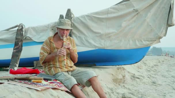 Senior man spelen bamboefluit op het strand naast vissersboot — Stockvideo