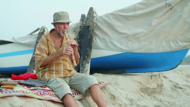 Senior man spelen bamboefluit op het strand naast vissersboot — Stockvideo