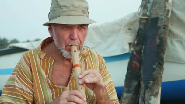 Portret van senior man bamboefluit spelen op het strand naast vissersboot — Stockvideo