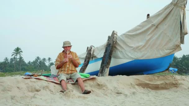 Senior man spelen bamboefluit op het strand naast vissersboot — Stockvideo