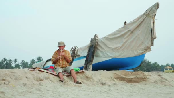 Homem sênior tocando flauta de bambu na praia ao lado do barco de pesca — Vídeo de Stock