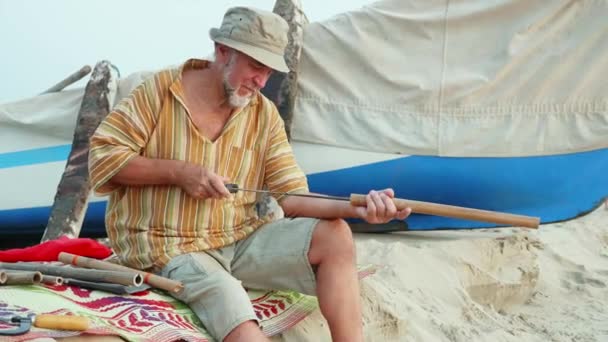 Hombre mayor se sienta en la playa y hace flauta con las manos . — Vídeos de Stock