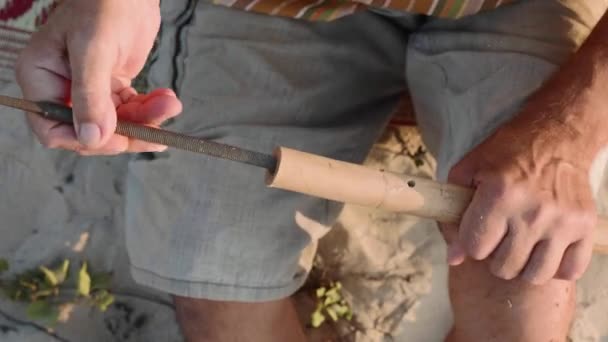 Senior man sits on the beach and making flute by hands, close-up. — Stock Video