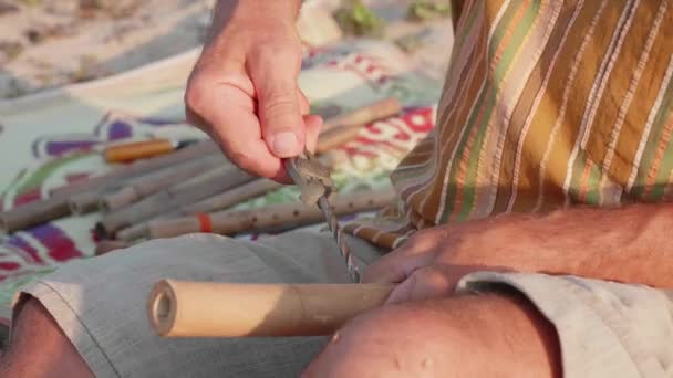 Senior man sits on the beach and making flute by hands, close-up. — Stock Video