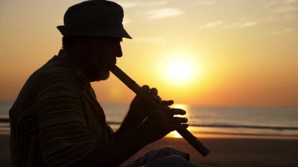 Sziluettjét magas rangú ember, bambusz fuvolán játszik a tengerparton naplementekor — Stock videók