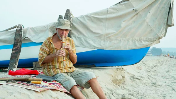 Homem sênior tocando flauta de bambu na praia ao lado do barco de pesca — Fotografia de Stock