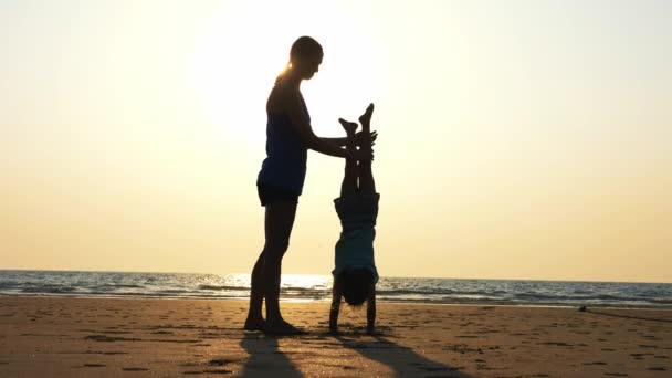 Силуэт матери с дочерью, занимающейся гимнастикой на пляже на закате — стоковое видео