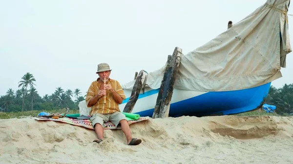 Старший играет на бамбуковой флейте на пляже рядом с рыбацкой лодкой — стоковое фото