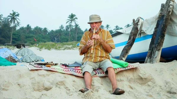 Homem sênior tocando flauta de bambu na praia ao lado do barco de pesca — Fotografia de Stock