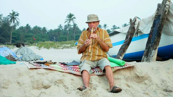 Senior man playing bamboo flute on the beach next to fishing boat