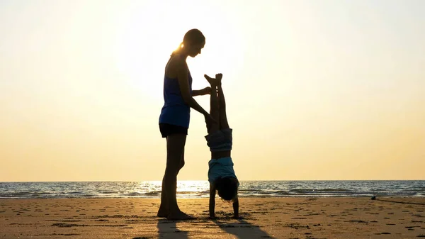 Silueta de madre con hija haciendo gimnasia en la playa al atardecer — Foto de Stock